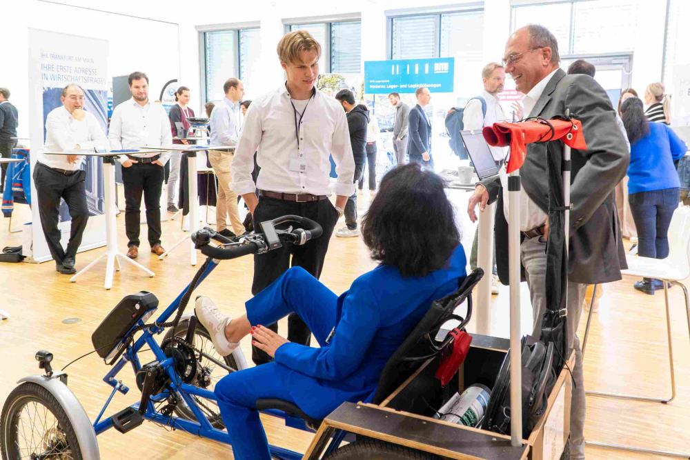 Eine Frau sitzt auf dem Prototypen eines Fahrzeugs mit vier Rädern und Pedalantrieb. Dabei ist sie im Austausch mit dem Start-up, die den Prototypen entwickelt haben. Im Hintergrund sind weitere Aussteller und Besucher zu sehen.