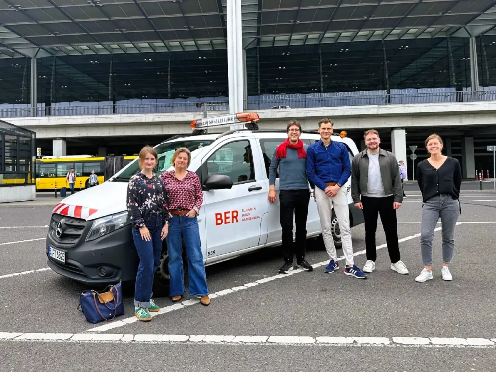 Gruppenbild mit sechs Personen. Die drei Frauen und drei Männer bilden das Projektteam des Förderprojets MoveBER, das von der HOLM GmbH, dem Flughafen Berlin-Brandenburg und der Universität Stuttgart von Mai 2026 bis Juni 2027 durchgeführt wird. Die Aufnahme entstand auf einem Parkplatz vor dem Flughafenterminal. Neben dem Gebäude sind ein Einsatzfahrzeug des Flughafens und ein Linienbus zu sehen.