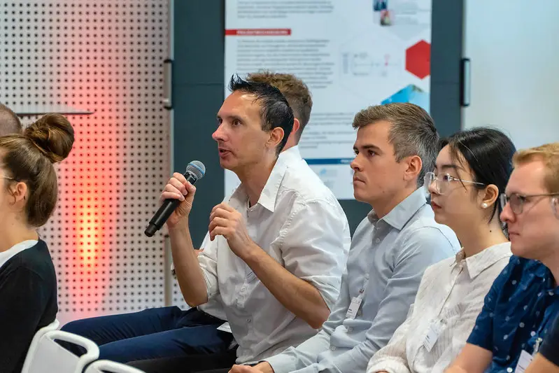Publikumsbild. Ein Mann hält ein Mikrofon in der Hand und beteiligt sich an der Diskussion. Im Hintergrund ist ein wissenschaftliches Projektposter zu sehen.